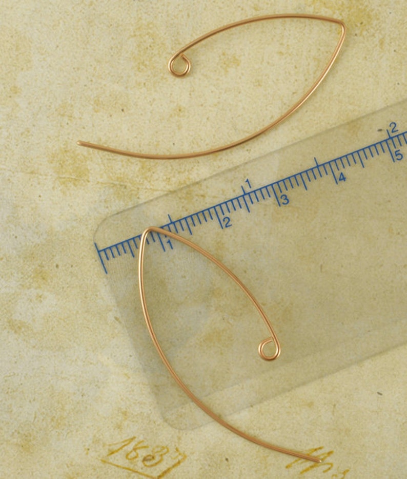May include: Two curved, gold-toned wire earring components with a loop at one end. A clear plastic ruler is positioned alongside the lower earring, showing a length of approximately 1.5 inches from the pointed tip to the curved base.