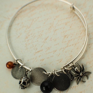 Puede incluir: Pulsera extensible plateada con m&uacute;ltiples dijes. Los dijes incluyen una calavera, una ara&ntilde;a, discos met&aacute;licos redondos y cuentas en negro, marr&oacute;n y plateado. La pulsera se muestra sobre un fondo con escritura.
