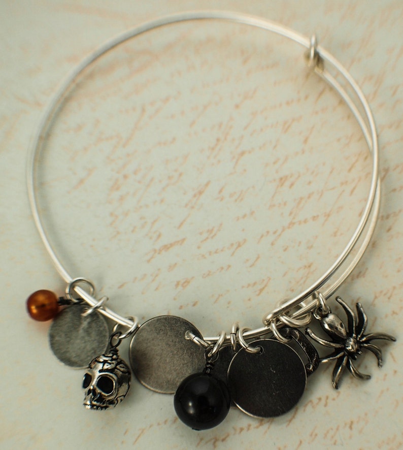Puede incluir: Pulsera extensible plateada con m&uacute;ltiples dijes. Los dijes incluyen una calavera, una ara&ntilde;a, discos met&aacute;licos redondos y cuentas en negro, marr&oacute;n y plateado. La pulsera se muestra sobre un fondo con escritura.