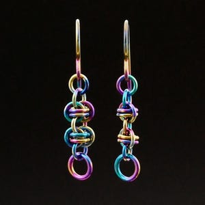 May include: A pair of iridescent earrings with a chainmail design. The earrings feature a series of colorful, interlocking rings in shades of purple, blue, green, and gold. The earrings hang from a simple hook.