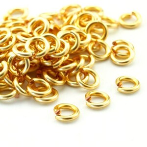 May include: A large collection of small, circular gold-colored metal jump rings. Each individual ring features a single open cut, revealing a copper-toned interior at the point of the opening. The rings are scattered in a pile on a white surface.
