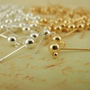 May include: A close-up of earring findings, some are silver and some are gold. The findings are on a light brown surface.