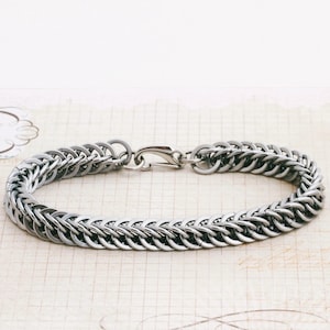 Könnte beinhalten: Ein silbernes Kettenarmband mit einem Karabinerverschluss. Die Kette besteht aus kleinen, ineinandergreifenden Ringen.