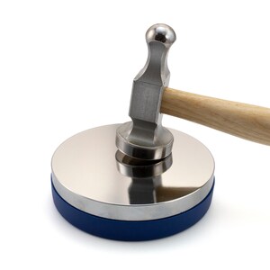 May include: A metal forming set with a polished steel disc and a ball-peen hammer. The disc has a blue base and the hammer has a wooden handle. The tools are designed for shaping and forming metal.