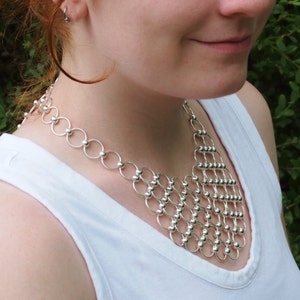 May include: A silver chain necklace with a geometric design. The necklace is made of small, round beads that are connected by thin, silver rings.