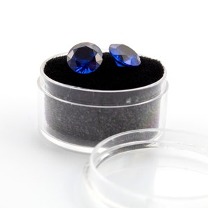 May include: Two dark blue round gemstones in a clear plastic container with a black velvet lining.