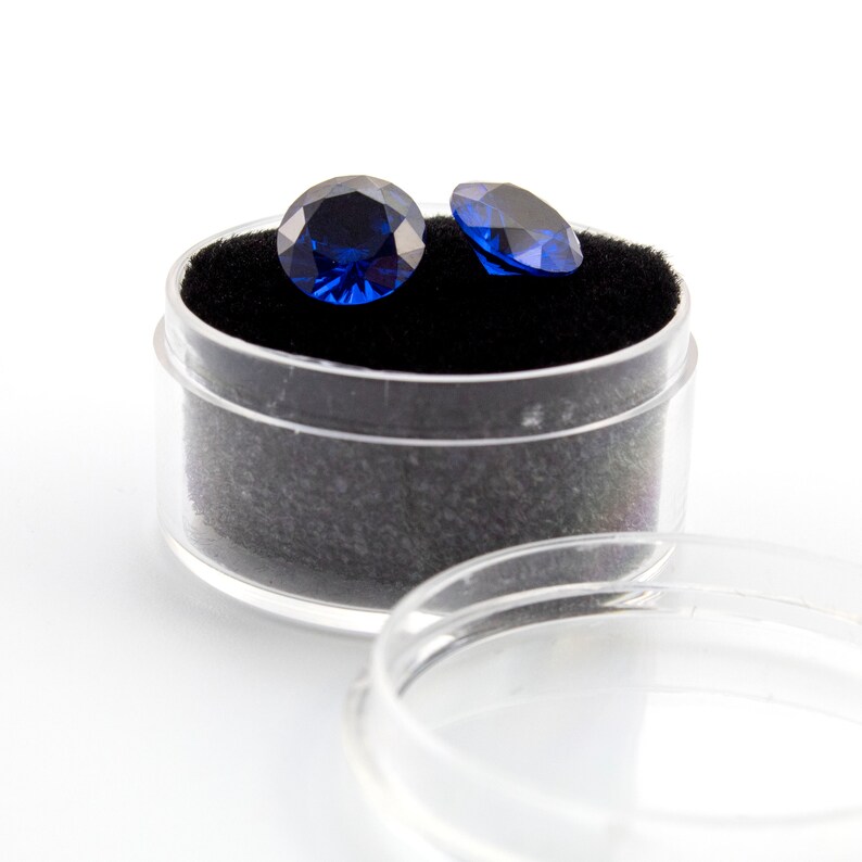 May include: Two dark blue round gemstones in a clear plastic container with a black velvet lining.