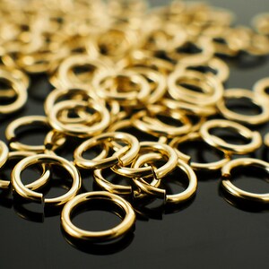 Op de afbeelding: Een verzameling glanzende, goudkleurige sprongringen, verspreid over een donker oppervlak. De ringen zijn open, klaar voor gebruik bij het maken van sieraden of andere ambachten. De close-up toont het gladde, reflecterende oppervlak van het metaal.