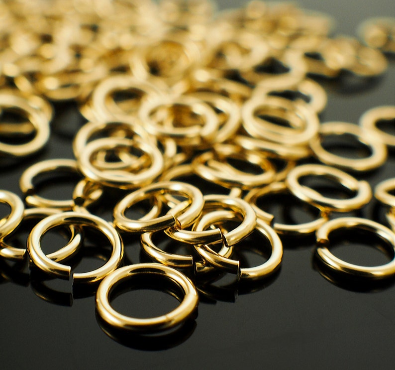Op de afbeelding: Een verzameling glanzende, goudkleurige sprongringen, verspreid over een donker oppervlak. De ringen zijn open, klaar voor gebruik bij het maken van sieraden of andere ambachten. De close-up toont het gladde, reflecterende oppervlak van het metaal.