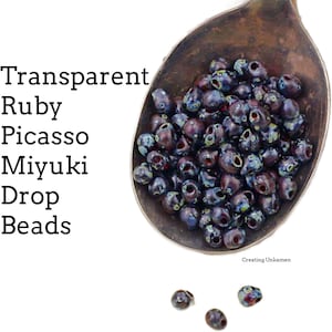May include: A close-up of a spoon filled with dark blue and red glass beads. The beads are small and have a speckled, iridescent finish. The text "Transparent Ruby Picasso Miyuki Drop Beads" is visible in the image.