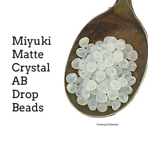 May include: A close-up of a spoon filled with white Miyuki Matte Crystal AB Drop Beads. The beads have a subtle iridescent sheen.