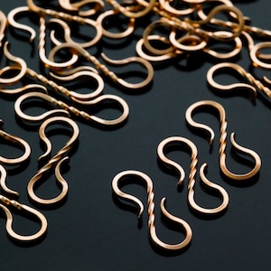 May include: A collection of small, gold-colored S-shaped clasps. Some clasps have a twisted design in the center, while others are smooth. The clasps are scattered on a dark surface, creating a visually appealing arrangement.
