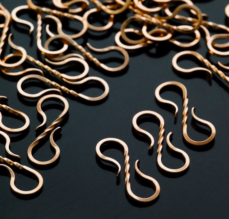 May include: A collection of small, gold-colored S-shaped clasps. Some clasps have a twisted design in the center, while others are smooth. The clasps are scattered on a dark surface, creating a visually appealing arrangement.