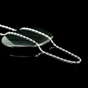 Könnte beinhalten: Eine zarte Silberkette mit ovalem Gliederdesign. Die Kette wird auf einer dunklen, reflektierenden Oberfläche vor schwarzem Hintergrund präsentiert. Die Halskette ist ein schlichtes, elegantes Schmuckstück.
