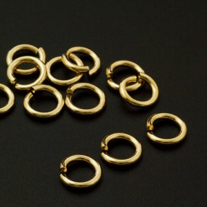 May include: A collection of small, circular gold-colored jump rings, each featuring a single open cut along the circumference. These metallic jewelry findings are arranged in a scattered pattern, showcasing a smooth, reflective surface finish.