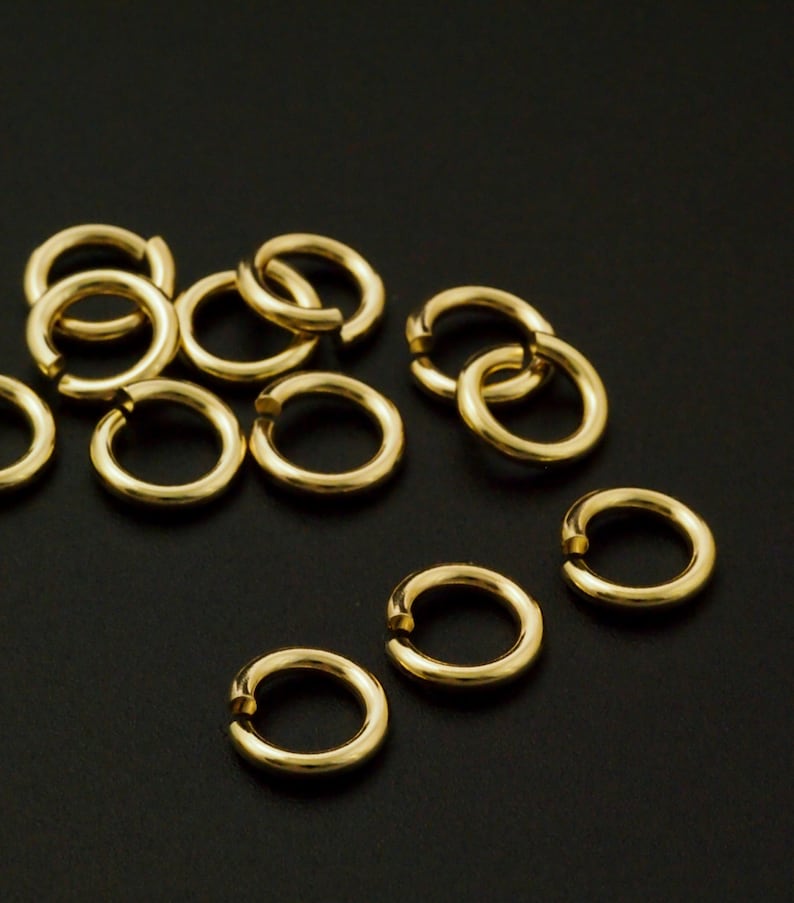 May include: A collection of small, circular gold-colored jump rings, each featuring a single open cut along the circumference. These metallic jewelry findings are arranged in a scattered pattern, showcasing a smooth, reflective surface finish.
