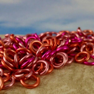 K&ouml;nnte beinhalten: Eine Ansammlung kleiner, kreisf&ouml;rmiger Metall-Biegeringe in verschiedenen Rosa-, Magenta- und Kupfert&ouml;nen. Jeder Ring weist eine einzelne Schnitt&ouml;ffnung auf, und das Sortiment ist in einem dichten, &uuml;berlappenden Haufen mit metallischem Glanz angeordnet.