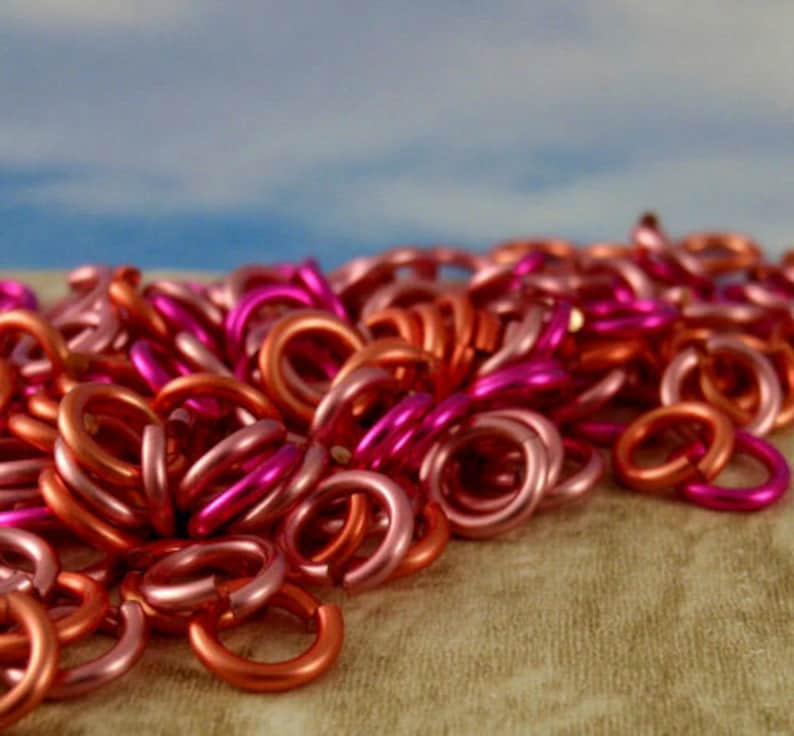 K&ouml;nnte beinhalten: Eine Ansammlung kleiner, kreisf&ouml;rmiger Metall-Biegeringe in verschiedenen Rosa-, Magenta- und Kupfert&ouml;nen. Jeder Ring weist eine einzelne Schnitt&ouml;ffnung auf, und das Sortiment ist in einem dichten, &uuml;berlappenden Haufen mit metallischem Glanz angeordnet.