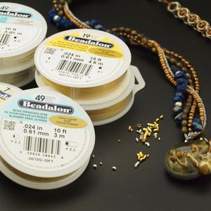 May include: Three round plastic spools of Beadalon bead stringing wire in gold and silver colors, each labeled with specifications including 0.024 inch diameter and lengths of 10 feet or 15 feet, alongside a multi-strand beaded necklace and small metal crimp beads.