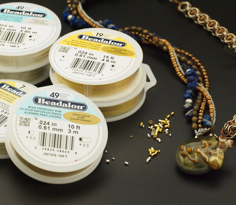 May include: Three round plastic spools of Beadalon bead stringing wire in gold and silver colors, each labeled with specifications including 0.024 inch diameter and lengths of 10 feet or 15 feet, alongside a multi-strand beaded necklace and small metal crimp beads.