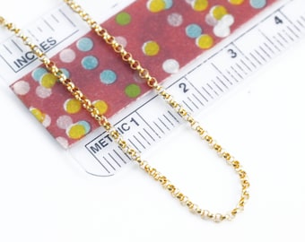 May include: A gold rolo chain necklace is positioned diagonally across a measuring tool. The ruler shows the chain length extending past 10 cm, with the links clearly visible against the white background and the colourful confetti pattern.