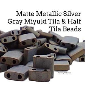 Op de afbeelding: Een stapel matte metallic zilvergrijze Miyuki Tila en halve Tila kralen. De kralen zijn rechthoekig met twee gaten in elk. De kralen zijn in een willekeurig patroon gerangschikt.