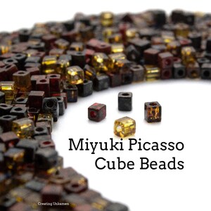May include: A close-up of a pile of small, colorful, square-shaped beads. The beads are in shades of red, brown, gold, and black. The text "Miyuki Picasso Cube Beads" is written below the beads.