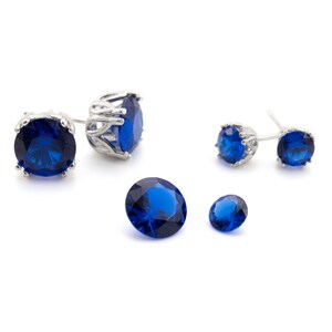 May include: Four blue sapphire earrings with silver settings. Two earrings have a larger stone and a more elaborate setting. The other two earrings have a smaller stone and a simpler setting.