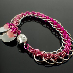 Pode incluir: Um bracelete de malha de corrente rosa e prata com um amuleto de prata e contas cor-de-rosa.