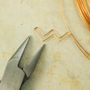May include: Close-up of a pair of silver pliers holding a small, zig-zag shaped piece of gold wire. The wire is on a light brown surface.