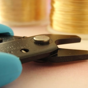 Puede incluir: Primer plano de unas pinzas para joyería negras y azules con una cabeza de metal plateado. Las pinzas están sobre una superficie rosa.