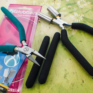 May include: Two pairs of jewelry pliers with black rubber grips. One pair has green handles and the other pair has silver handles. The pliers are on a pink and white background with the text "Wubbers Made to Make Jewelry" visible.