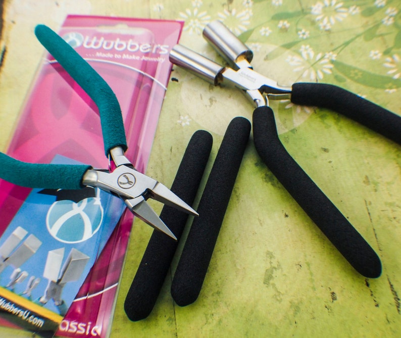 May include: Two pairs of jewelry pliers with black rubber grips. One pair has green handles and the other pair has silver handles. The pliers are on a pink and white background with the text "Wubbers Made to Make Jewelry" visible.