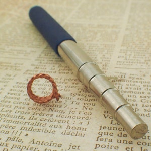 Puede incluir: Primer plano de un bolígrafo telescópico retráctil azul y plateado sobre un trozo de papel con texto. El bolígrafo está extendido y la punta es visible.