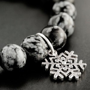 May include: A bracelet composed of round, polished snowflake obsidian beads featuring a black and white mottled pattern. A silver-toned metal charm shaped like a six-pointed snowflake is attached to the bracelet by a small circular jump ring.