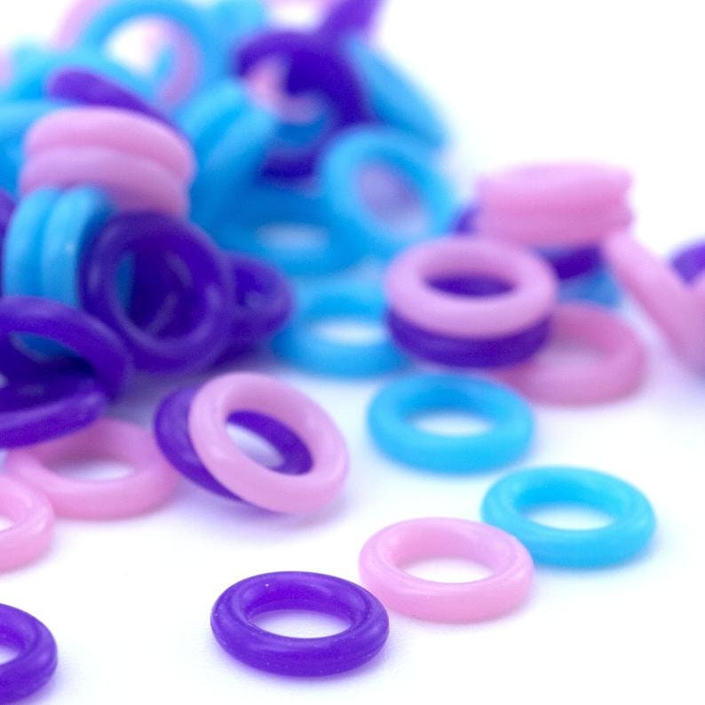 K&ouml;nnte beinhalten: Ein Haufen bunter Plastikringe in Rosa, Blau und Lila. Die Ringe sind klein und rund und liegen auf einer wei&szlig;en Oberfl&auml;che.