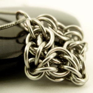Puede incluir: Un collar de cota de malla en tono plateado que presenta una serie de anillos met&aacute;licos circulares entrelazados. La cadena est&aacute; construida con un patr&oacute;n tejido denso que crea una superficie met&aacute;lica texturizada. Una cadena de serpiente fina est&aacute; unida al lado.