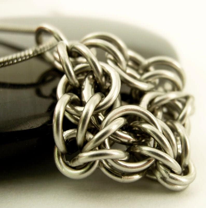 Puede incluir: Un collar de cota de malla en tono plateado que presenta una serie de anillos met&aacute;licos circulares entrelazados. La cadena est&aacute; construida con un patr&oacute;n tejido denso que crea una superficie met&aacute;lica texturizada. Una cadena de serpiente fina est&aacute; unida al lado.