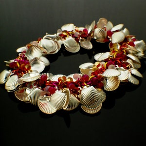 K&ouml;nnte beinhalten: Ein Bettelarmband mit zahlreichen silberfarbenen Muschel-Anh&auml;ngern, durchsetzt mit kleinen, runden, durchscheinenden Perlen in Dunkelrosa, Hellrosa und Bernsteint&ouml;nen, die alle durch eine Reihe von metallischen Ringen in Pink und Gold verbunden sind.