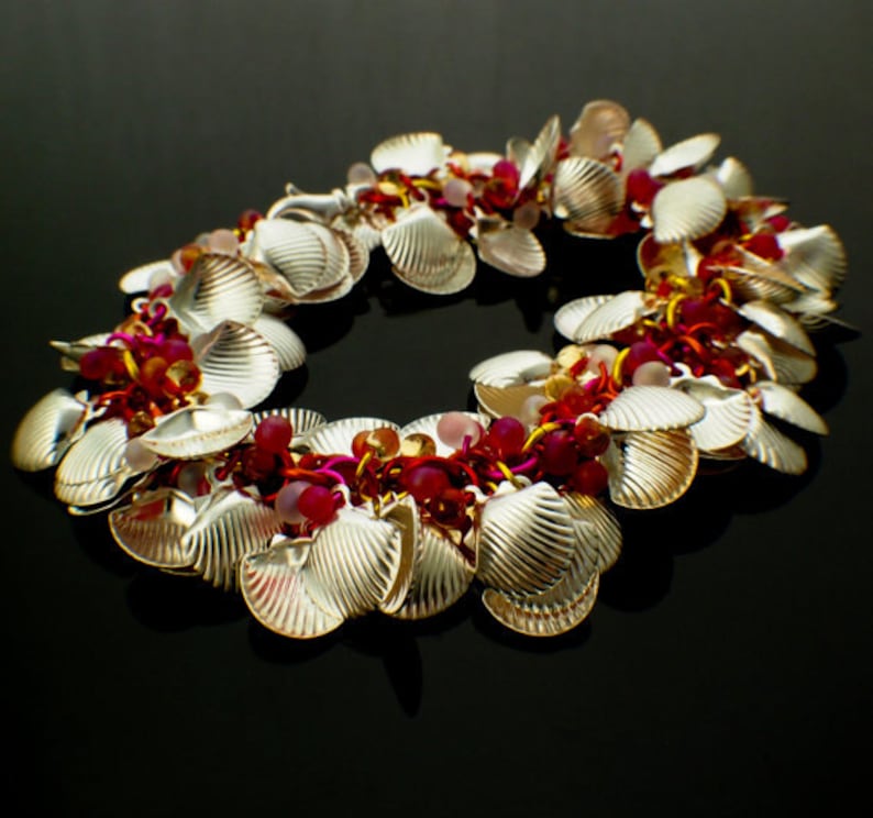 K&ouml;nnte beinhalten: Ein Bettelarmband mit zahlreichen silberfarbenen Muschel-Anh&auml;ngern, durchsetzt mit kleinen, runden, durchscheinenden Perlen in Dunkelrosa, Hellrosa und Bernsteint&ouml;nen, die alle durch eine Reihe von metallischen Ringen in Pink und Gold verbunden sind.