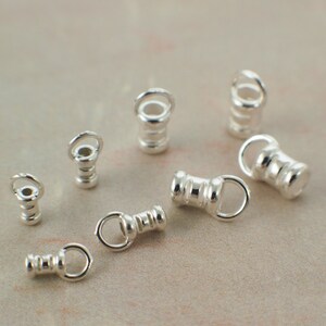 May include: A collection of small, silver-colored jewelry findings. The findings have a cylindrical shape with a loop at one end and a small ring at the other. They are scattered on a neutral-toned surface.