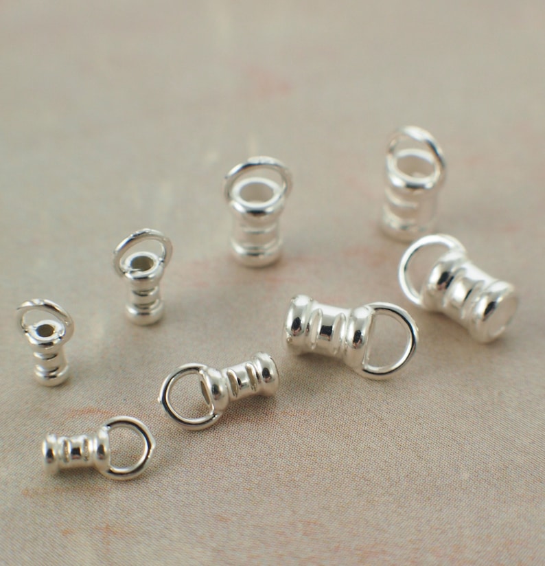 May include: A collection of small, silver-colored jewelry findings. The findings have a cylindrical shape with a loop at one end and a small ring at the other. They are scattered on a neutral-toned surface.