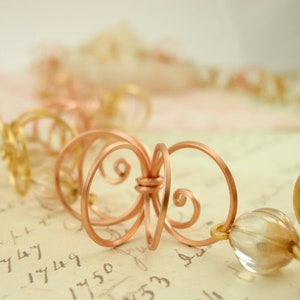 May include: A copper wire necklace with a delicate, intricate design. The necklace features a series of small, interconnected loops and swirls, creating a flowing, organic pattern. The necklace is accented with clear glass beads.