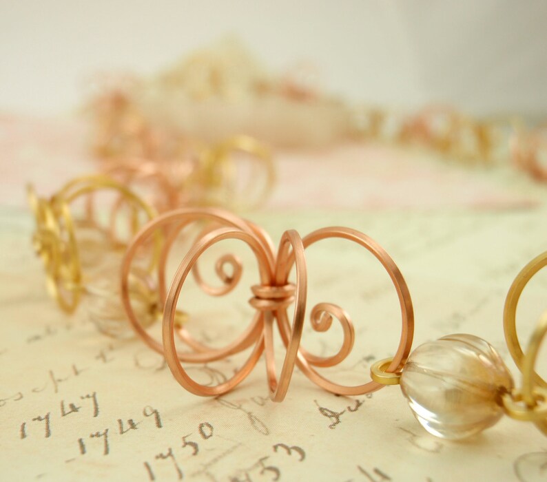 May include: A copper wire necklace with a delicate, intricate design. The necklace features a series of small, interconnected loops and swirls, creating a flowing, organic pattern. The necklace is accented with clear glass beads.