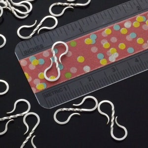 May include: Close-up of several silver-colored S-shaped clasps, some with a twisted design, scattered on a black surface. A ruler with inch measurements and a colorful dotted pattern is in the background. The clasps are approximately 1 inch in length.