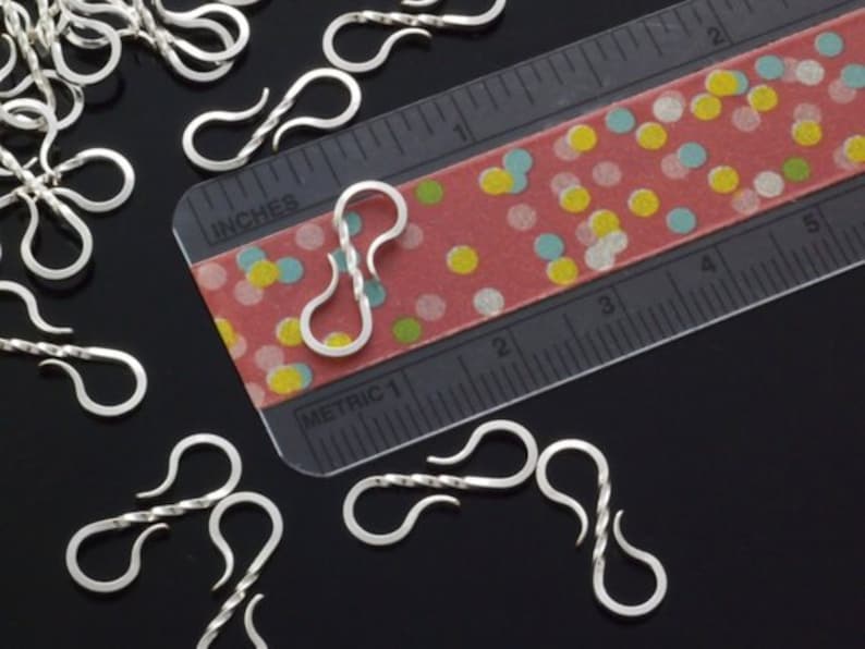 May include: Close-up of several silver-colored S-shaped clasps, some with a twisted design, scattered on a black surface. A ruler with inch measurements and a colorful dotted pattern is in the background. The clasps are approximately 1 inch in length.