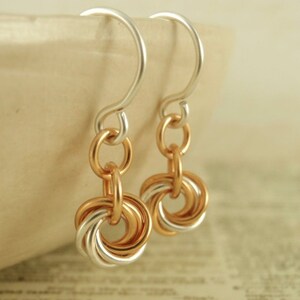 May include: A pair of drop earrings featuring silver-toned ear wires connected to a series of interlocking circular rings in alternating rose gold and silver tones. The design consists of multiple small, thin metal loops linked together to form a circular pendant shape that hangs from the curved ear wire.