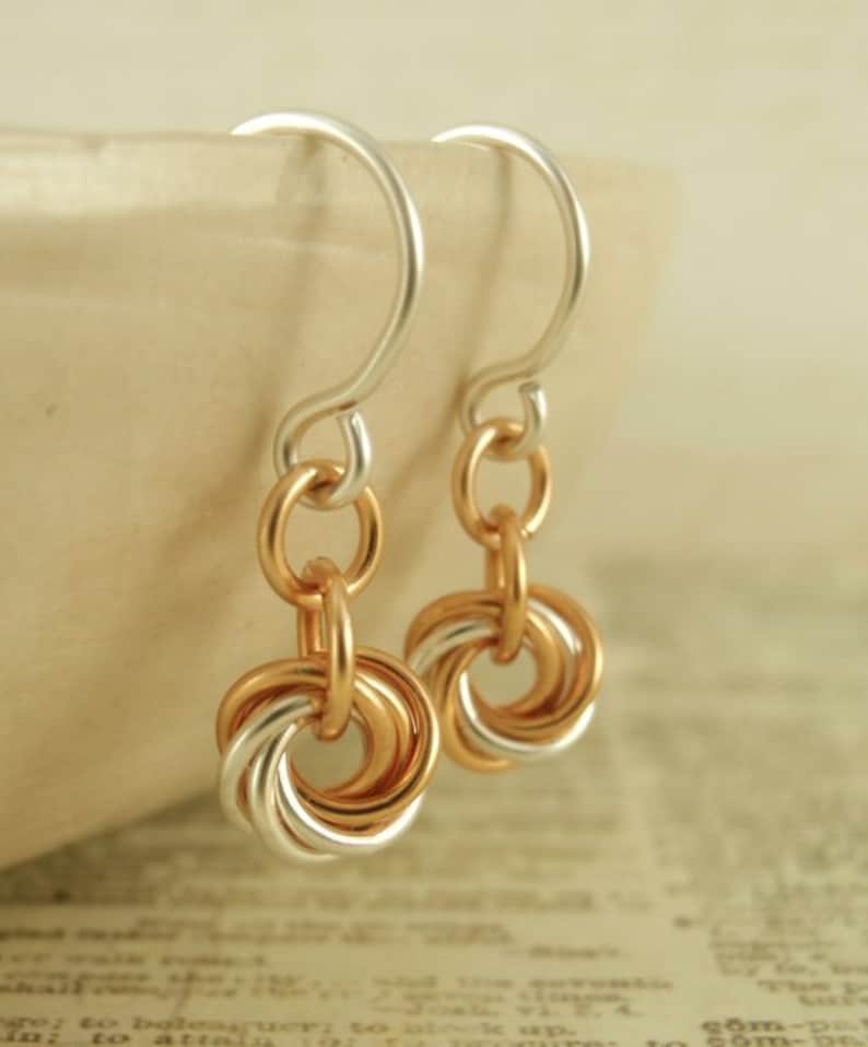 May include: A pair of drop earrings featuring silver-toned ear wires connected to a series of interlocking circular rings in alternating rose gold and silver tones. The design consists of multiple small, thin metal loops linked together to form a circular pendant shape that hangs from the curved ear wire.