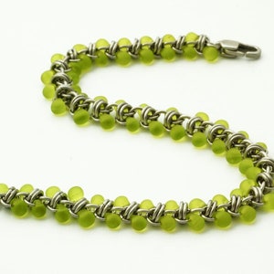 Puede incluir: Una pulsera de cota de malla construida con anillos de metal plateado entrelazados y peque&ntilde;as cuentas redondas de color verde lima transl&uacute;cido. La pulsera presenta un patr&oacute;n repetitivo de eslabones met&aacute;licos y cuentas, con un cierre de mosquet&oacute;n plateado.