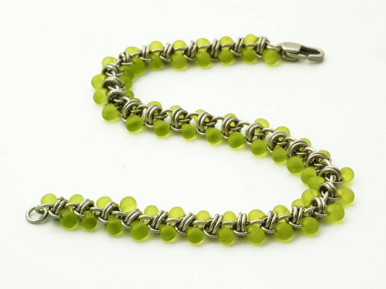 Puede incluir: Una pulsera de cota de malla construida con anillos de metal plateado entrelazados y peque&ntilde;as cuentas redondas de color verde lima transl&uacute;cido. La pulsera presenta un patr&oacute;n repetitivo de eslabones met&aacute;licos y cuentas, con un cierre de mosquet&oacute;n plateado.